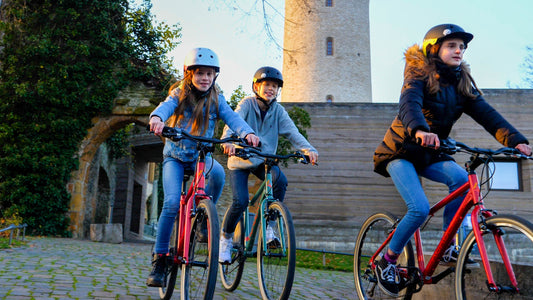 Drei Teenager fahren auf ihren Academy Kinderfahrrädern durch einen Park. Hinter ihnen ist ein steinerner Turm