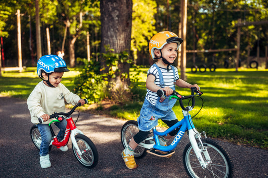Zwei Kinder fahren auf einem roten und einem blauen Woom Kinderfahrrad durch den Park