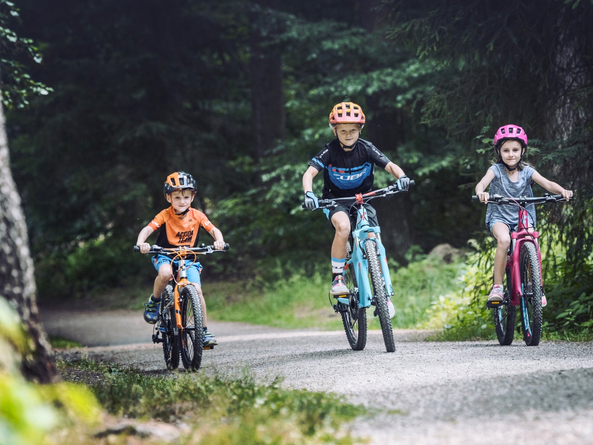 Drei Kinder in sportlicher Kleidung und Fahrradhelmen fahren auf ihren bunten Cube Kinderfahrrädern durch einen Pfad im Wald.