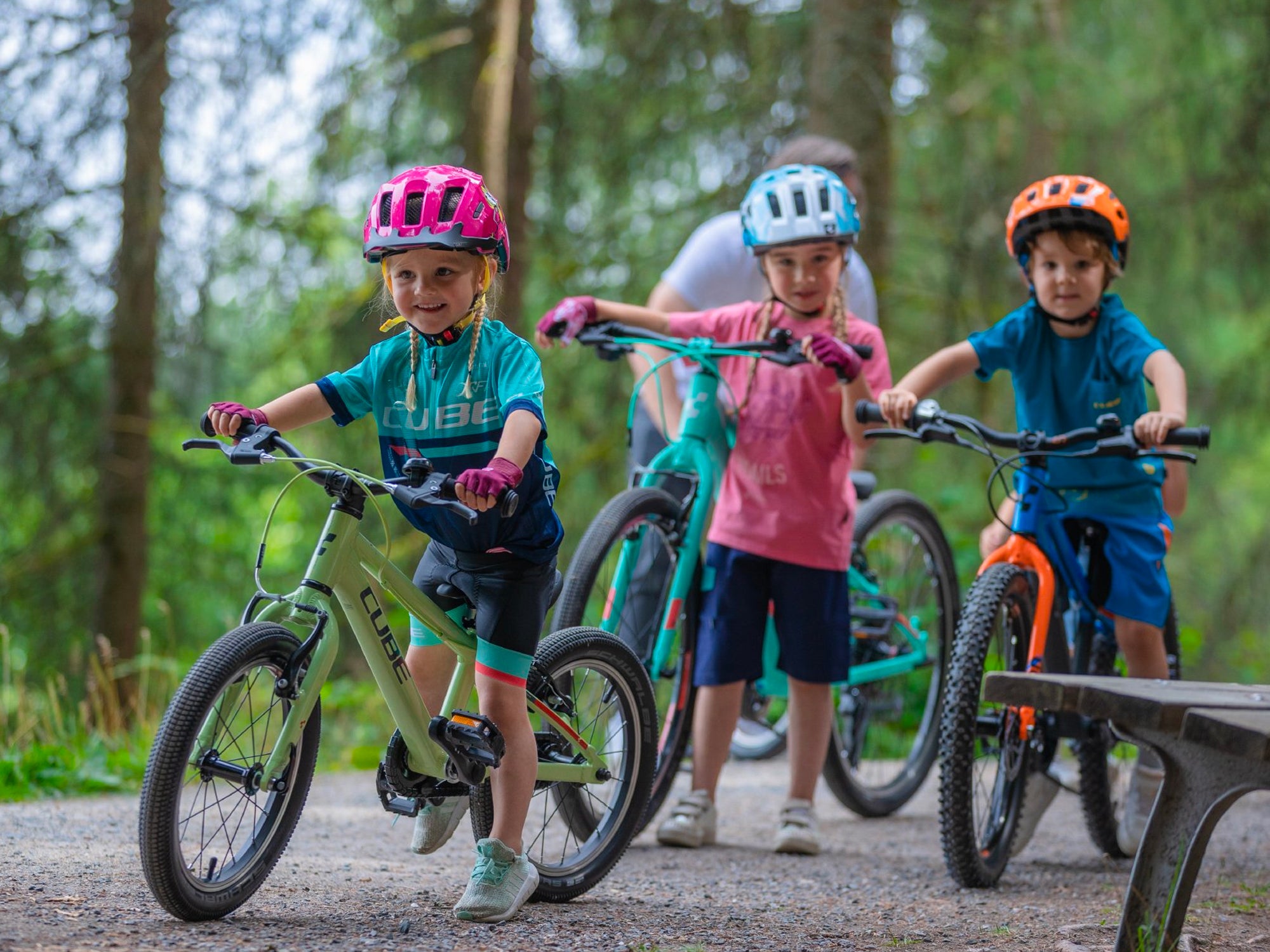 Drei junge Kinder in sportlicher Kleidung und Fahrradhelmen stehen mit ihren Cube Kinderfahrrädern auf einem Pfad in einem Wald.
