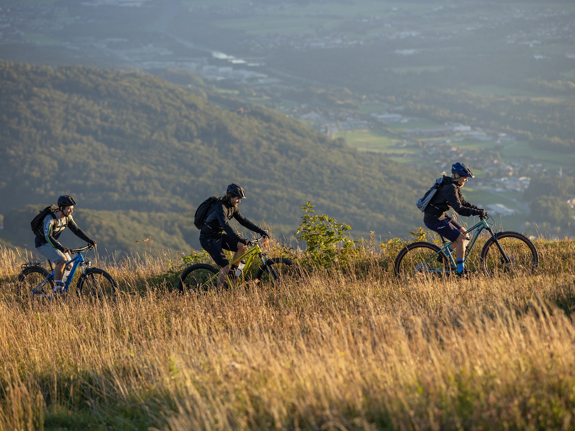 Drei Menschen in sportlicher Kleidung und Fahrradhelmen fahren auf ihren Cube Fahrrädern auf einem Naturpfad in einer hügeligen Landschaft.