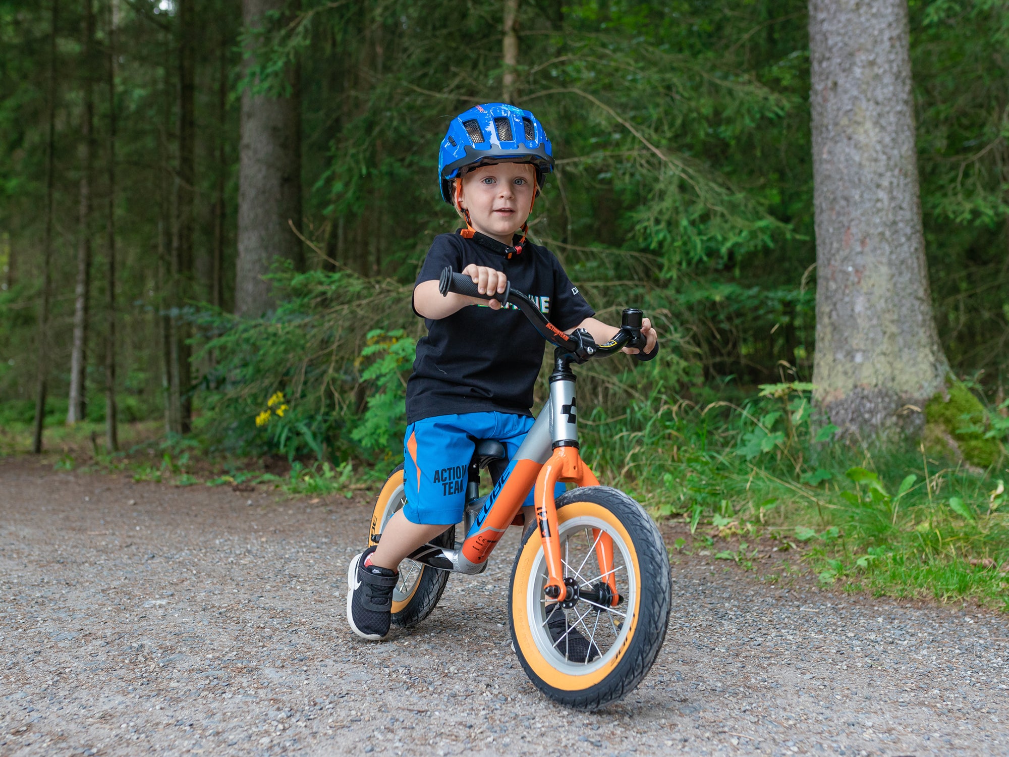 Ein kleiner Junge in sportlicher Kleidung und einem blauen Fahrradhelm fährt mit einem Cube Laufrad auch den Wald