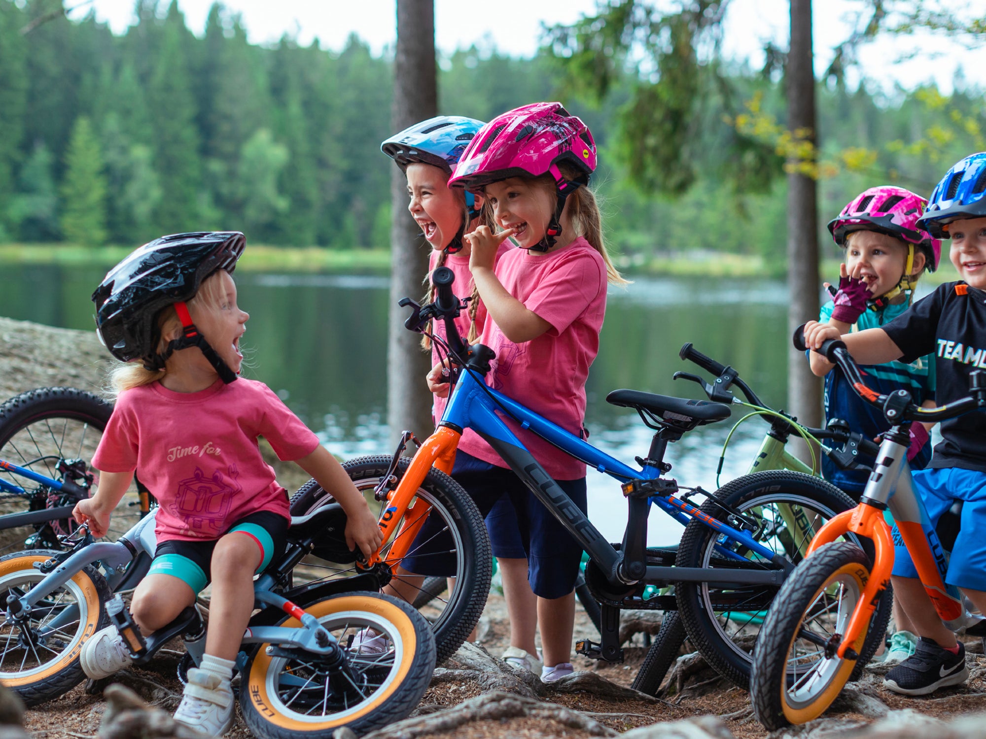 Fünf Kinder in sportlicher Kleidung und Fahrradhelmen stehen mit ihren Cube Kinderfahrrädern neben einem See in einem Wald
