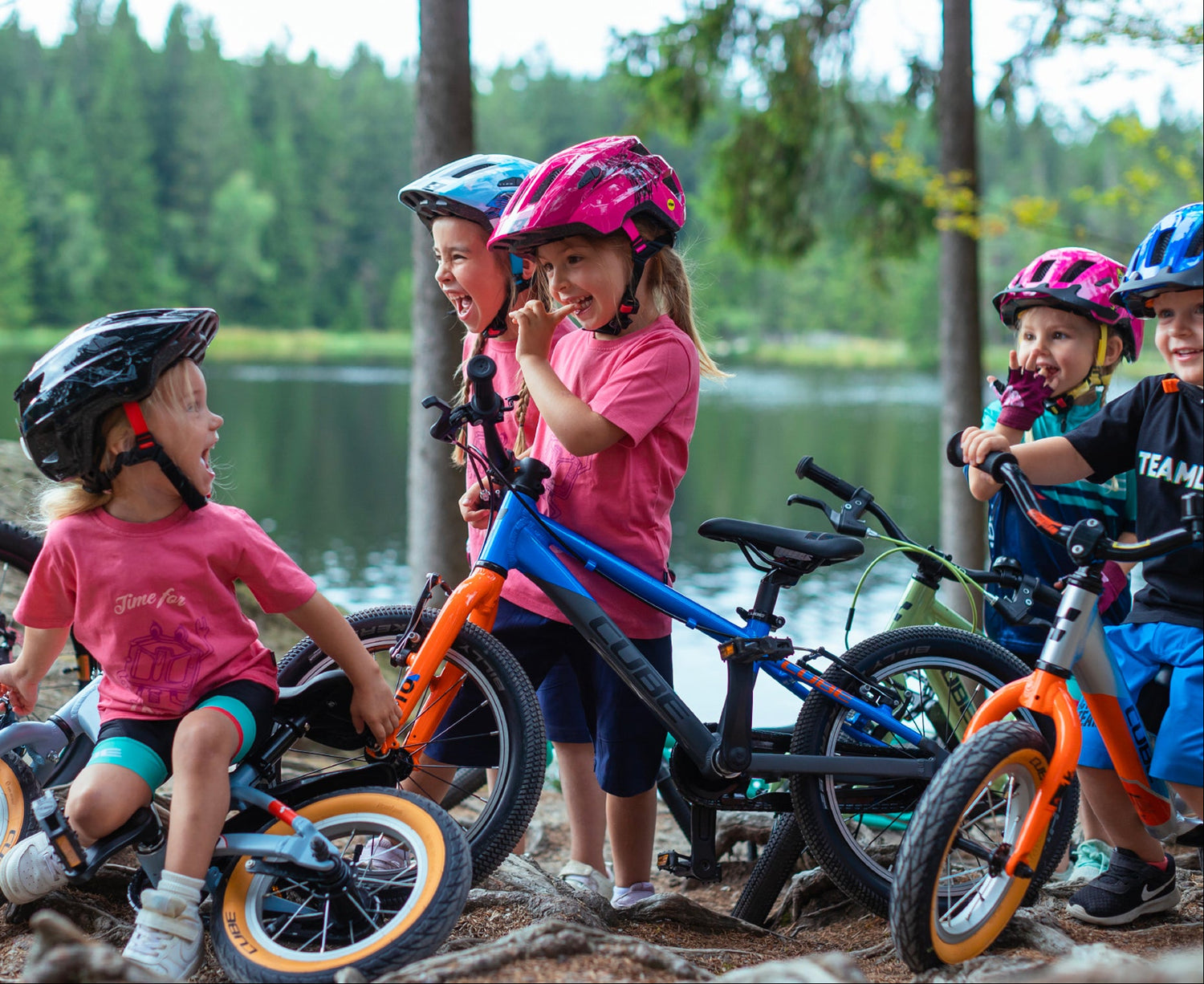 Fünf Kinder in sportlicher Kleidung und Fahrradhelmen stehen mit ihren Cube Kinderfahrrädern neben einem See in einem Wald